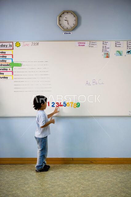 كتابة وعد الأرقام بالترتيب, تعلم الأعداد والأرقام الانجليزية, صورة من الخلف لطالب تمهيدي عربي خليجي إماراتي يرتدي الزي الكاجوال يقف أمام السبورة, حصص اللغة الإنجليزية في مدارس الإمارات العربية المتحدة, تطور طرق التعليم وتنمية الذكاء لدى الأولاد