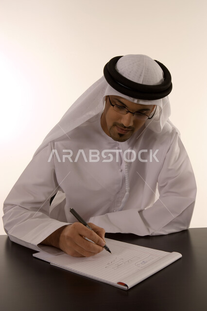ايماءات التركيز على العمل, انجاز المهام وتحقيق الاهداف, تدوين الملاحظات ومراجعتها, ملفات ووثائق خاصة بالعمل, بورتريه لرجل خليجي عربي اماراتي يرتدي الكندورة الاماراتية والعمامة يقوم بالعمل داخل مكتبه, خلفية بيضاء