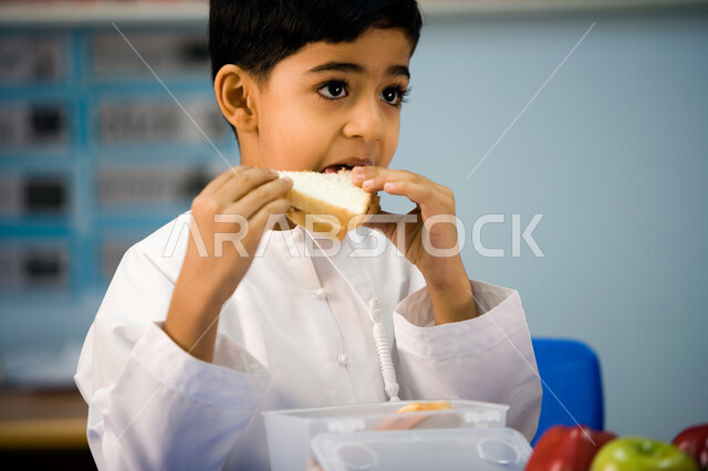 تعليم أطفال التمهيدي أسس الغذاء الصحي السليم, ايماءات الاستمتاع بتناول الشطيرة الصحية، صورة مقربة لطفل عربي خليجي اماراتي مبتسم يرتدي الزي الاماراتي ينظر إلى شيء ما, تناول الأطعمة الصحية في وقت الفسحة المدرسية في إحدى المدارس الاماراتية