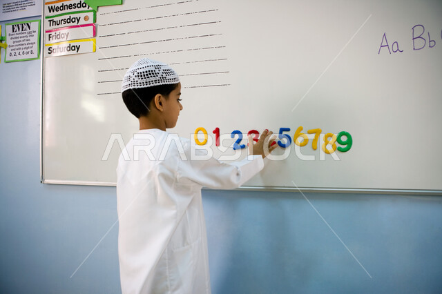 حصص اللغة الانجليزية في مدارس الإمارات العربية المتحدة, صورة من الخلف لتلميذ عربي خليجي إماراتي يرتدي الكندورة والطاقية البيضاء يكتب على السبورة, تعلم عد وكتابة الأرقام بالأنجليزي, الاهتمام بالتعليم وتقوية المهارات الحركية لأطفال التمهيدي