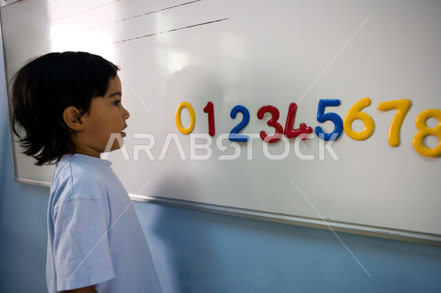 تعلم الأرقام والأعداد الانجليزية, صورة مقربة من الجانب لطالب تمهيدي عربي خليجي إماراتي يرتدي الزي الكاجوال يقف أمام السبورة, حصص اللغة الإنجليزية في مدارس الإمارات العربية المتحدة, تطور طرق التعليم وتنمية الذكاء لدى الأولاد 