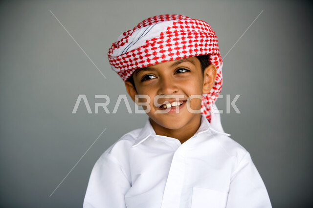 التخطيط والتفكير بالأهداف المستقبلية, النظر إلى شيء ما بإيماءات السرور والسعادة, بورتريه مقرب لفتى عربي خليجي إماراتي مبتسم يرتدي الزي الإماراتي التقليدي ينظر للأعلى, الاستمتاع بقضاء وقت ممتع, خلفية رمادي