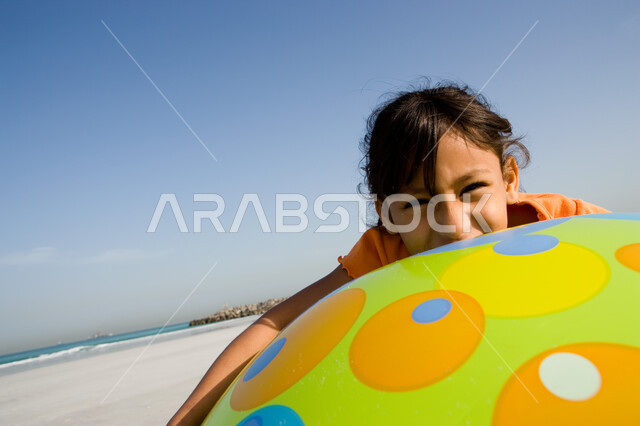 Gestures of happiness and enjoyment of spending the summer vacation on the sea, beach resorts in the UAE, a close-up of a smiling young Arab Emirati Gulf girl holding her hand in a large rubber ball on the beach, entertaining leisure activities on the hol