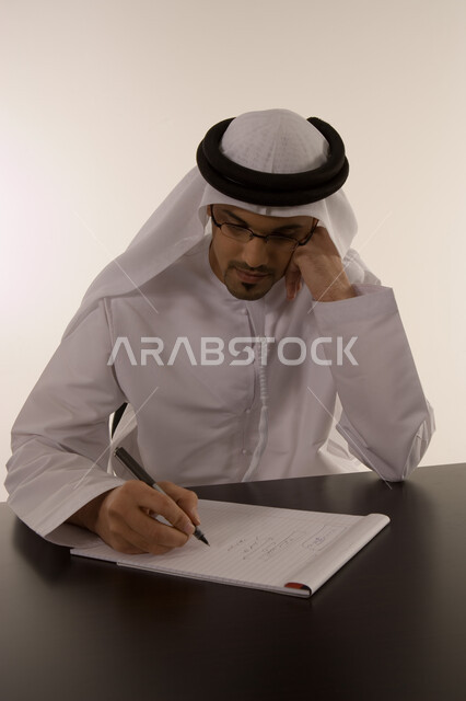 ايماءات التركيز على العمل, انجاز المهام وتحقيق الاهداف, تدوين الملاحظات ومراجعتها, ملفات ووثائق خاصة بالعمل, بورتريه لرجل خليجي عربي اماراتي يرتدي الكندورة الاماراتية والعمامة يقوم بالعمل داخل مكتبه, خلفية بيضاء