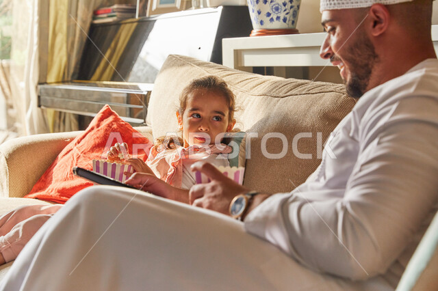  تبادل النظرات والاحاديث بين الفتاة ووالدها، متابعة المسلسلات والأفلام المسلية، اجواء اسرية مليئة بالسعادة و السرور، أب عربي خليجي اماراتي يرتدي الكندورة والطاقية البيضاء الاماراتيه يقوم بمشاهدة التلفاز وتناول الفوشار برفقة إبنته، قضاء اوقات عائلية ممتعة.