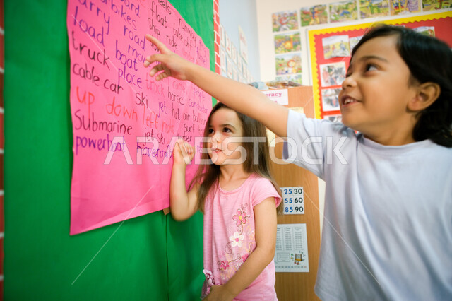 تعليم اللغات الأجنبية لطلاب المرحلة التمهيدية, الطريقة الصحيحة لقراءة الدرس، المدارس الأكاديمية في الإمارات العربية المتحدة، صورة مقربة لولد وبنت عربيين خليجيين إماراتيين يقفان أمام السبورة, الإشارة إلى النص الانجليزي ومحاولة القراءة