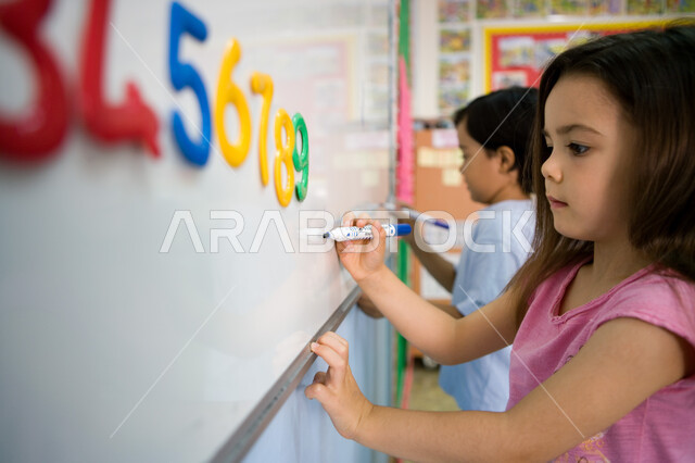 اتباع الطريقة الصحيحة لرسم وكتابة الأرقام, تعليم اللغات الأجنبية لطلاب المرحلة التمهيدية، المدارس الأكاديمية في الإمارات العربية المتحدة، صورة مقربة من الجانب لطالبين عربيين خليجين إماراتيين يكتبان أرقام اللغة الانجليزية على السبورة في الروضة