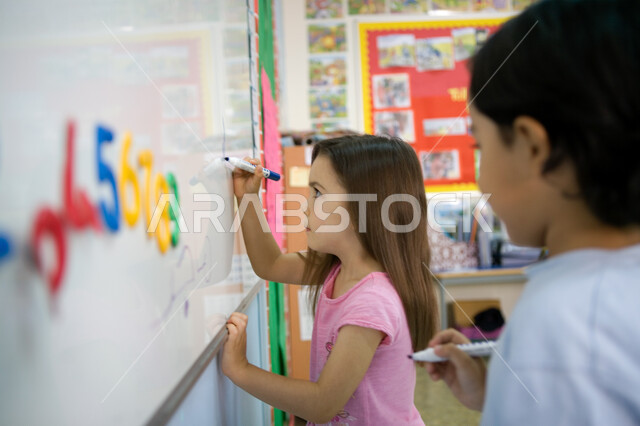 اتباع الطريقة الصحيحة لرسم وكتابة الأرقام, تعليم اللغات الأجنبية لطلاب المرحلة التمهيدية، المدارس الأكاديمية في الإمارات العربية المتحدة، صورة مقربة من الجانب لطالبين عربيين خليجين إماراتيين يكتبان أرقام اللغة الانجليزية على السبورة في الروضة