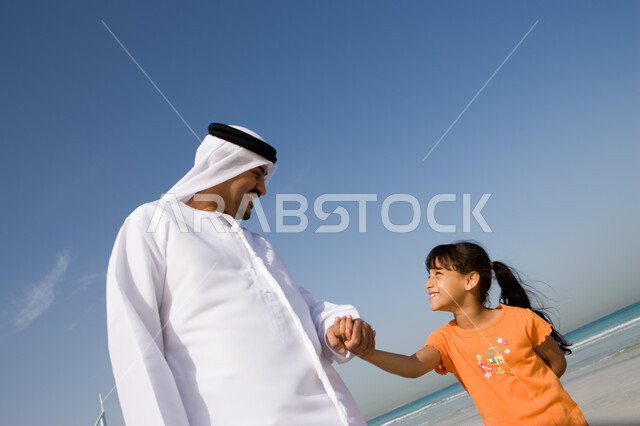 الاستمتاع والسعادة بالسير مع الأبناء على شاطئ البحر في الإجازة الصيفية, مفهوم الرابطة الأبوية, ايماءات الحب والعطف على الأبناء, صورة مقربة لأب عربي خليجي إماراتي يقوم بالنظر الى ابنته و يسير معها على الشاطئ,  القيام بالفعاليات الصيفية مع الأبناء