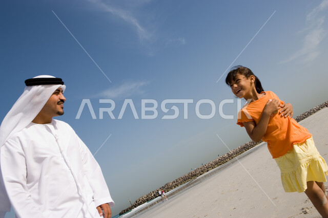 السير مع الأبناء على شاطئ البحر في الإجازة الصيفية, مفهوم الرابطة الأبوية, ايماءات الحب والعطف على الأبناء, صورة مقربة لأب عربي خليجي إماراتي يقوم بالنظر الى ابنته و يسير معها على الشاطئ,  القيام بالفعاليات الصيفية مع الأبناء