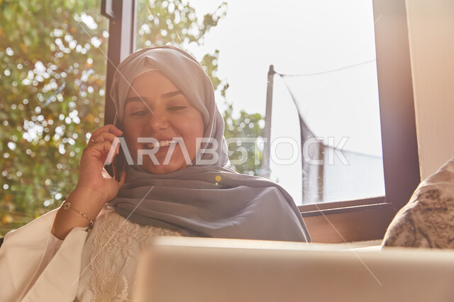 Facial gestures indicating happiness and pleasure in making a phone call, a veiled Arab Gulf Emirati woman sitting on the sofa talking on a mobile phone in broad daylight, looking at something, spending pleasant times exchanging news and conversations, co