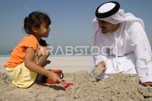 مشاركة الآباء الأبناء نشاطاتهم الترفيهية, قضاء أوقات صيفية ممتعة باللعب بالرمال على ساحل البحر, صورة مقربة لفتاة صغيرة عربية خليجية اماراتية تقوم باللعب بأدوات الرمل رفقة والدها على شاطئ البحر في إجازة الصيف، السياحة في دول الخليج العربي