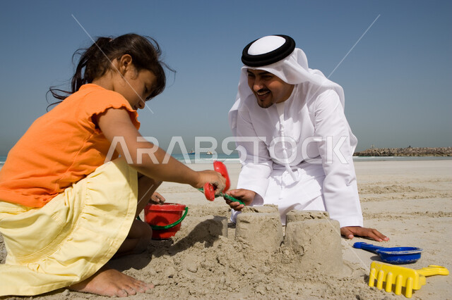 Parents sharing their recreational activities with their children, spending fun summer times playing with sand on the sea coast, a close-up of a young Arab Gulf Emirati girl playing with sand tools with her father on the seashore during the summer vacatio