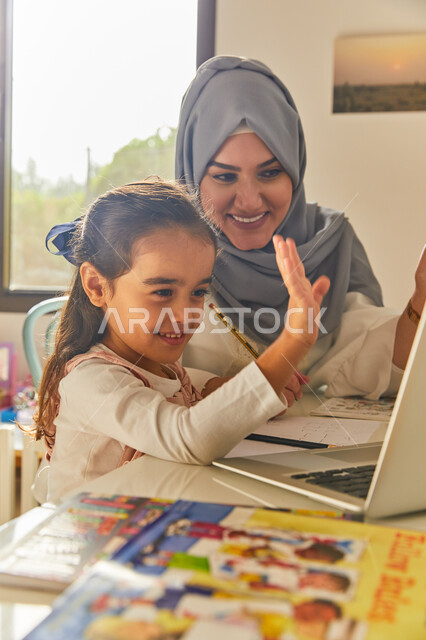 Making a video call via a laptop, showing a live broadcast on one of the social media, a veiled Arab Gulf Emirati mother who welcomes and communicates online with relatives and friends accompanied by her daughter, following the e-learning platforms availa