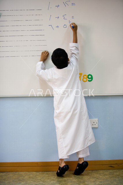 القيام بالعمليات الحسابية في مادة الرياضيات، جودة التعليم في الإمارات العربية المتحدة، صورة من الخلف لطالب عربي خليجي إماراتي يرتدي الزي الاماراتي يقوم بحل التمارين على السبورة في الروضة، تنمية القدرات الفكرية ومهارات الحساب لطلاب المرحلة التمهيدية