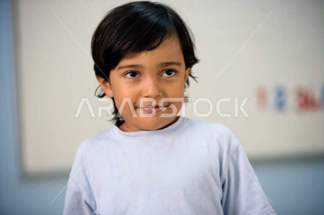 Looking at something with gestures of pleasure, a close-up of an Arab Gulf Emirati child in the preparatory stage wearing casual clothes standing in the classroom, absent-mindedness and thinking about an answer, education, innovation and the development o