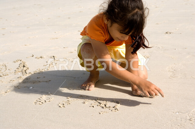 التسلية والترفيه بقضاء أوقات صيفية ممتعة, الاستمتاع باللعب بالرمال على ساحل البحر, صورة مقربة لفتاة صغيرة عربية خليجية اماراتية تقوم بالرسم والكتابة على شاطئ البحر في إجازة الصيف، السياحة في دول الخليج العربي