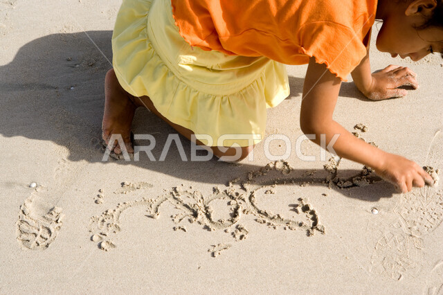 التسلية والترفيه بقضاء أوقات صيفية ممتعة, الاستمتاع باللعب بالرمال على ساحل البحر, صورة مقربة لفتاة صغيرة عربية خليجية اماراتية تقوم بالرسم والكتابة على شاطئ البحر في إجازة الصيف، السياحة في دول الخليج العربي