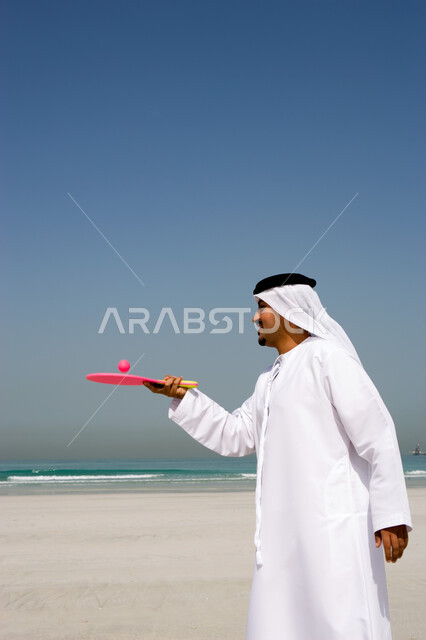 Spending vacation times with entertainment and enjoyment, entertaining with interesting summer activities, recreational activities on vacation, an Emirati Arab man wearing a ghutra and an Emirati kandora playing tennis on the beach, beach resorts in the E