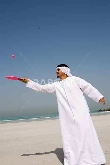 قضاء أوقات الإجازة بالترفيه والاستمتاع, التسلية  بالفعاليات الصيفية الممتعة , أنشطة ترفيهية  في العطلة, رجل عربي خليجي إماراتي يرتدي الغترة والكندورة الإماراتية يقوم باللعب بكرة المضرب على شاطئ البحر, منتجعات شاطئية في الإمارات