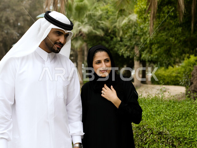 Exchanging conversations and news, an Arab Gulf Emirati couple walking and wandering in the open air, hiking during vacation time in one of the Emirates parks, leisure activities and spending fun family times, the green nature background.
