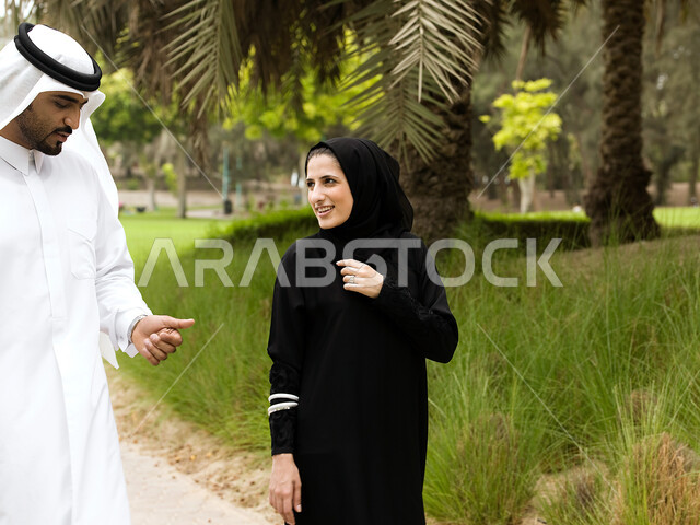 تبادل الأحاديث والأخبار, زوجان عربيان خليجيان إماراتيان يقومان بالمشي و التجول في الهواء الطلق, التنزه في وقت العطلة في إحدى حدائق الإمارات, أنشطة ترفيهية وقضاء أوقات عائلية ممتعة, خلفية الطبيعة الخضراء.