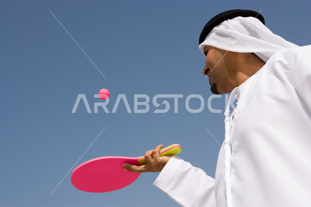 اللعب بكرة التنس في العطلة, قضاء أوقات الإجازة بالترفيه والاستمتاع, التسلية  بالفعاليات الصيفية الممتعة , صورة مقربة لرجل عربي خليجي إماراتي يرتدي الغترة والكندورة الإماراتية يقوم باللعب بكرة المضرب على شاطئ البحر, منتجعات شاطئية في الإمارات
