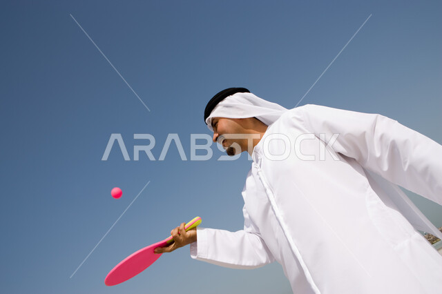 اللعب بكرة التنس في العطلة, قضاء أوقات الإجازة بالترفيه والاستمتاع, التسلية  بالفعاليات الصيفية الممتعة , صورة مقربة لرجل عربي خليجي إماراتي يرتدي الغترة والكندورة الإماراتية يقوم باللعب بكرة المضرب على شاطئ البحر, منتجعات شاطئية في الإمارات