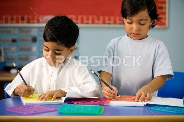 ايماءات تدل على الاندماج والتركيز لطلاب التمهيدي, تعلم مهارة الدقة والابتكار لدى الطلاب، صورة مقربة لطالبين عربيين خليجين اماراتيين يرتديان الزي الكاجوال الاماراتي ويمسكان أدوات الرسم، التعلم عن طريق اللعب داخل الفصل