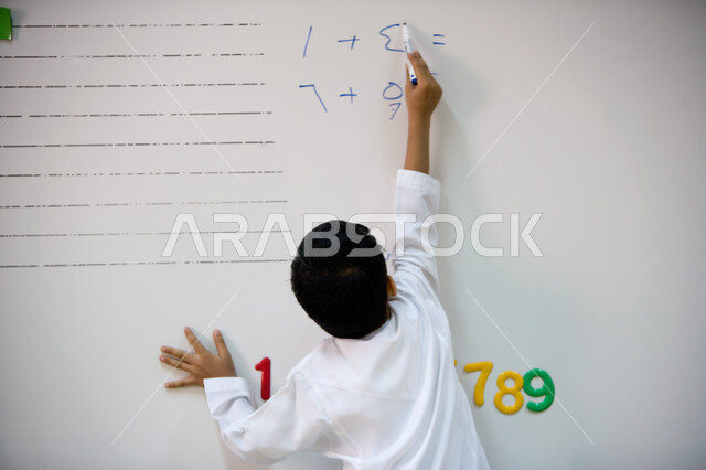 Learning arithmetic operations in mathematics, the quality of education and schools in the United Arab Emirates, a back image of an Arab Gulf Emirati student wearing Emirati uniforms doing exercises on the board in kindergarten, developing intellectual ab