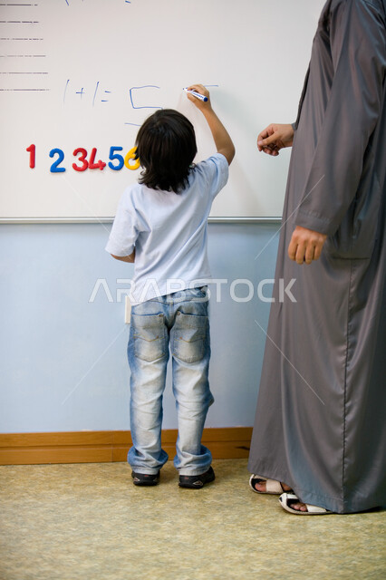 المشاركة العملية ومحاكاة ذكاء الطالب, التعلم وتنمية المهارات الذهنية لدى الأطفال, صورة من الخلف لطالب عربي خليجي إماراتي يقوم بالكتابة على السبورة داخل الفصل برفقة معلمته, القيام بالعمليات الحسابية, الأساليب الحديثة في التعليم الاستنتاجي