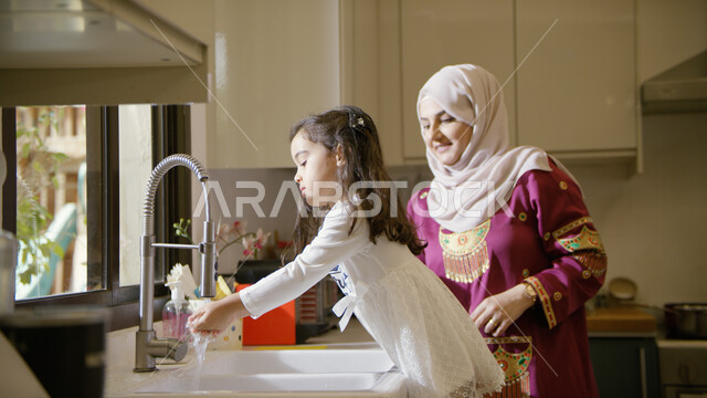 Educating children about the need to adhere to the teachings of personal hygiene in the time of corona, an Arab Emirati Gulf woman wearing an embroidered abaya standing next to her daughter, washing hands with soap and water to follow methods of care and 