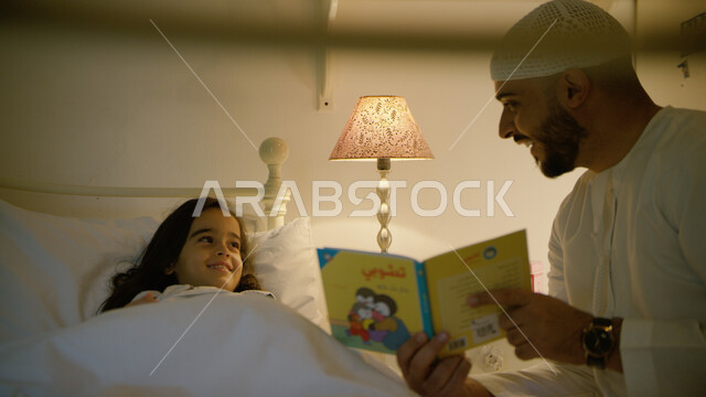 انشطة ترفيهيه قبل النوم, مشاركه الأبناء بقراءه القصص المفيده قبل النوم، مفهوم التربيه القائمه على الحب والحنان، رجل عربي خليجي إماراتي يرتدي الكندوره والطاقيه البيضاء الاماراتيه يقرأ الروايات المسليه لابنته، قضاء وقت ممتع مع الأبنه
