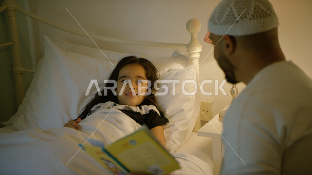 مفهوم التربيه على الحب والحنان، انشطة ترفيهيه قبل النوم, مشاركه الأبناء بقراءه القصص المفيده قبل النوم، رجل عربي خليجي إماراتي يرتدي الكندوره والطاقيه البيضاء الاماراتيه يقرأ الروايات المسليه لابنته، قضاء وقت ممتع مع الأبنه