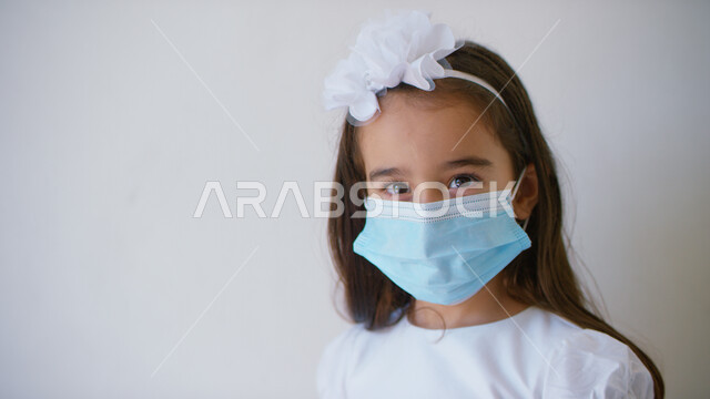 The concept of following preventive measures to ensure safety from the Corona virus, health awareness and avoiding the spread of infection, a close-up portrait of an Arab Gulf Emirati girl wearing a white dress and wearing a muzzle to protect from infecti