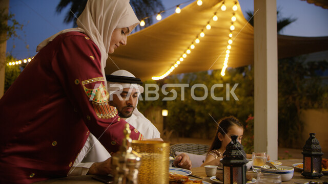 قدوم شهر الخير والبركة, المأكولات والمقبلات الرمضانيه المتنوعه والشهية، اجتماع الأهل ومشاركة الطعام على مائده الإفطار في شهر رمضان المبارك، امرأة عربيه خليجيه اماراتية تقوم بصب الطعام لزوجها وابنتها في الخارج، لأضواء والديكورات الرمضانية  .