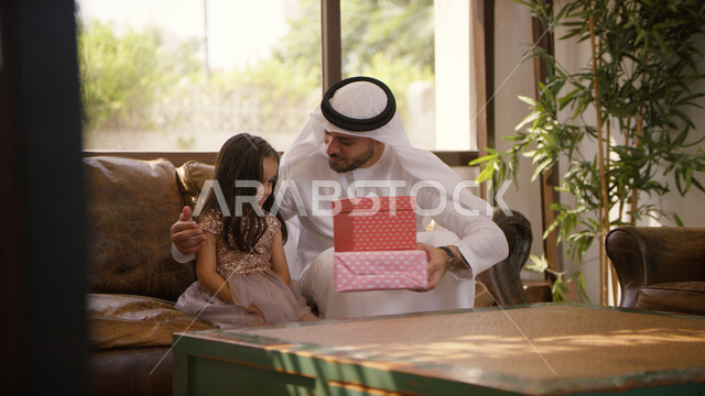 Exchanging conversations and chats with children, congratulating on the advent of the blessed Eid, a close-up of an Arab, Emirati father wearing an Emirati kandora and ghutra sitting in the living room presenting gifts to his daughter on holidays and occa