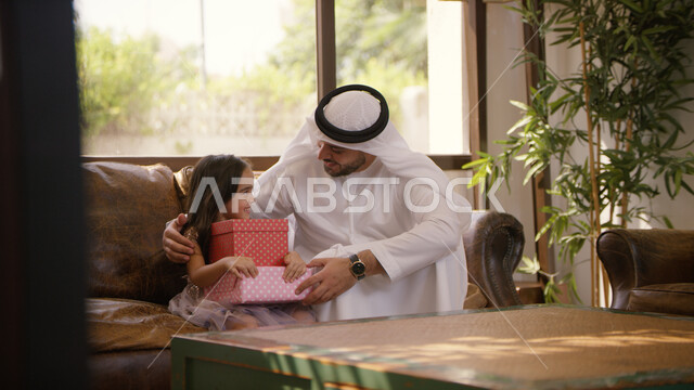  الاحتفال بقدوم العيد السعيد, أجواء عائلية مليئة بالحب، ايماءات الفرح بتقديم الهدايا في الأعياد والمناسبات الإسلامية، صورة مقربه لرجل عربي اماراتي خليجي يرتدي الكندورة و الغترة الاماراتية يجلس في غرفة المعيشة مع ابنته، الضحك والتسامر مع الأبناء