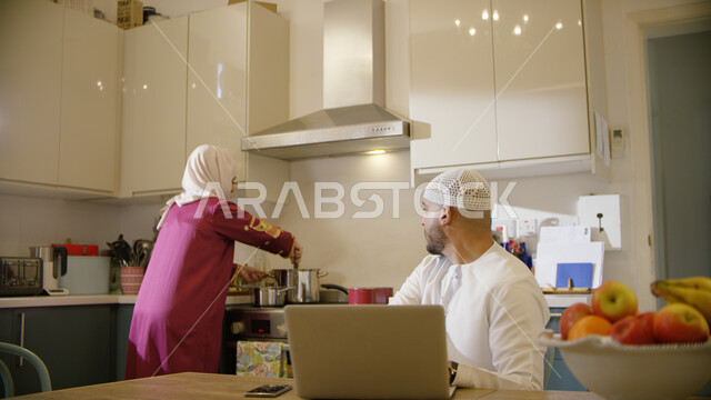 Exchanging conversations between the spouses while cooking food, making a voice call via mobile phone, spending quality time in the kitchen with an Emirati Arab man wearing a kandora and a cap working on a laptop, a veiled Emirati Arab Gulf woman wearing 