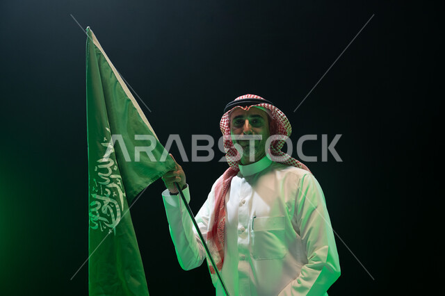 The concept of love for the homeland and belonging to it, celebrating the anniversary of the Saudi National Day, September 23, a close-up portrait of a smiling Gulf Arab man wearing a thobe and shemagh and holding the Kingdom’s flag in his hand with gestures of pride and pride in the homeland, Saudi Flag Day, March 11, the Saudi National Day, black background