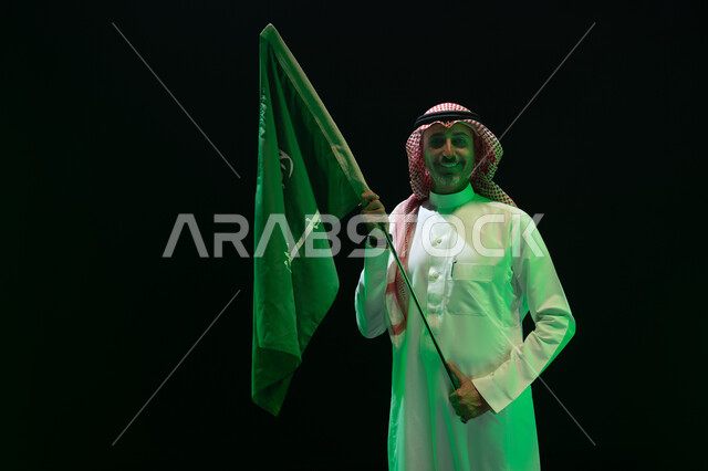 Commemoration of Saudi National Day, September 23, celebration of Saudi National Day, portrait of a smiling Saudi Gulf Arab man wearing the Saudi thobe and shemagh and holding the flag of the Kingdom of Saudi Arabia in his hand, Saudi Flag Day, March 11, Saudi National Day, black background