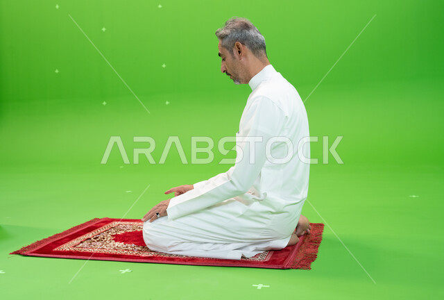 الجلوس للتشهد على سجادة الصلاة, الخشوع في الصلاة والتضرع الى الله ، كروما لرجل عربي خليجي سعودي يرتدي الزي التقليدي يجلس على سجادة الصلاة ،العبادة والتقرب إلى الله، مفهوم أركان الإسلام، اسلاميات وعبادة، خلفية خضراء