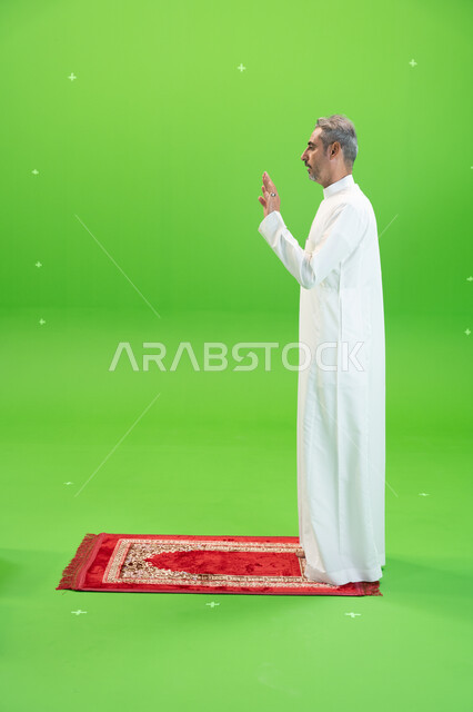 أداء فرائض الصلاة بأوقاتها، كروما لرجل عربي خليجي سعودي يرتدي الزي التقليدي يقف على سجادة الصلاة ويرفع يديه لتكبيرة الإحرام و الاستعداد للبدء بالصلاة ،العبادة والتقرب إلى الله، إسلاميات وعبادة، صورة الجسم بالطول الكامل ،خلفية خضراء