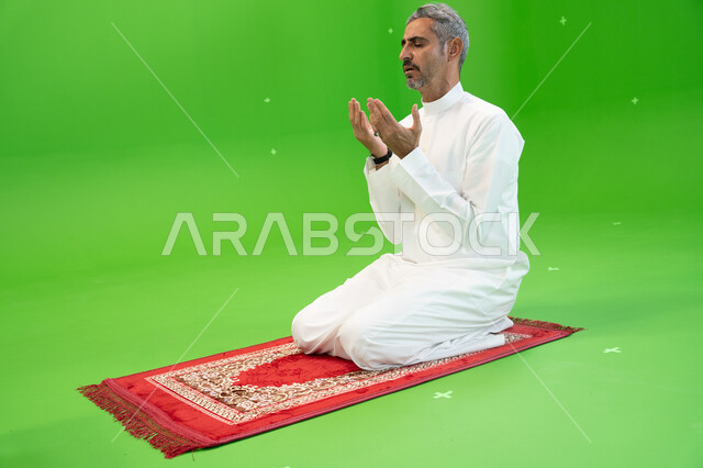 اسلاميات وعبادة، التقرب الى الله بالأعمال الصالحة، التذلل و الطلب من الله تعالى, كروما لرجل عربي خليجي سعودي يرتدي الزي التقليدي يجلس على سجادة الصلاة و يرفع يديه ويدعي بإيماءات التوسل و الخشوع، خلفية خضراء