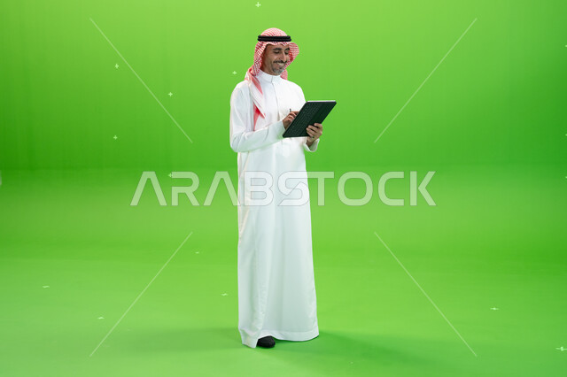 صورة الجسم بالطول الكامل، التسويق الإلكتروني عبر الأجهزة الحديثة، تصفح مواقع التواصل الإجتماعي، كروما لرجل عربي خليجي يرتدي الزي التقليدي السعودي ويحمل بيده جهاز التابليت ويقرأ عبارات فكاهية مضحكة، متابعه آخر أخبار السوشال ميديا، خلفية خضراء