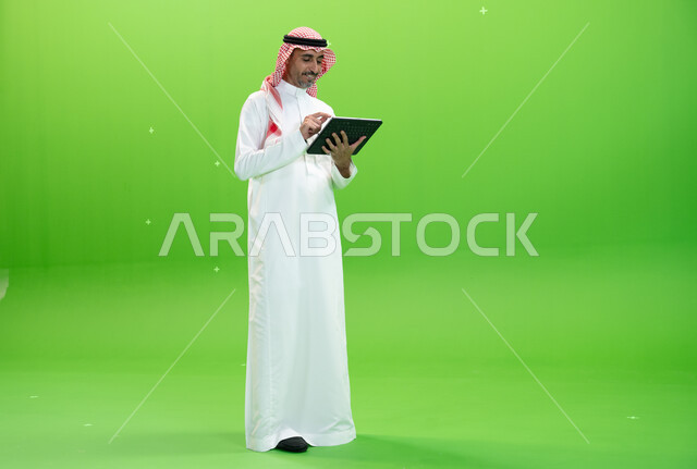 التسويق الإلكتروني عبر الأجهزة الحديثة، تصفح مواقع التواصل الإجتماعي، كروما لرجل عربي خليجي يرتدي الزي التقليدي السعودي ويحمل بيده جهاز التابليت، البحث عن عمل عن طريق الانترنت،متابعه آخر أخبار السوشال ميديا، صورة الجسم بالطول الكامل، خلفية خضراء