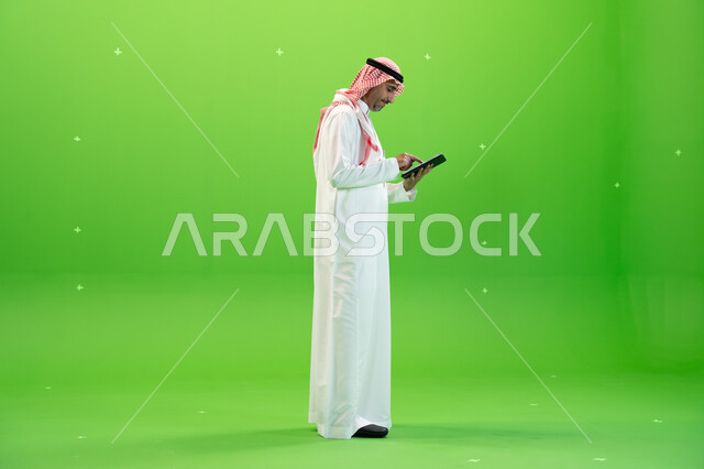 التسويق الإلكتروني عبر الأجهزة الحديثة، تصفح مواقع التواصل الإجتماعي، كروما لرجل عربي خليجي يرتدي الزي التقليدي السعودي ويحمل بيده جهاز التابليت، البحث عن عمل عن طريق الانترنت،متابعه آخر أخبار السوشال ميديا، صورة الجسم بالطول الكامل، خلفية خضراء