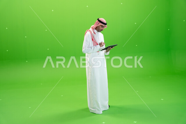 E-marketing through modern devices, browsing social networking sites, chroma of an Arab Gulf man wearing traditional Saudi dress and holding a tablet in his hand, searching for a job via the Internet, following the latest social media news, full-length bo
