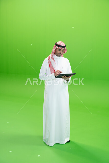 تصفح مواقع التواصل الإجتماعي، البحث عن عمل عن طريق الانترنت، كروما لرجل عربي خليجي يرتدي الزي التقليدي السعودي ويحمل بيده جهاز التابليت، التسويق الإلكتروني عبر الأجهزة الحديثة، متابعه آخر أخبار السوشال ميديا، صورة الجسم بالطول الكامل، خلفية خضراء