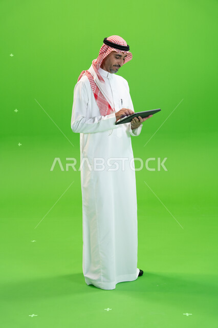 تصفح مواقع التواصل الإجتماعي، البحث عن عمل عن طريق الانترنت، كروما لرجل عربي خليجي يرتدي الزي التقليدي السعودي ويحمل بيده جهاز التابليت، التسويق الإلكتروني عبر الأجهزة الحديثة، متابعه آخر أخبار السوشال ميديا، صورة الجسم بالطول الكامل، خلفية خضراء